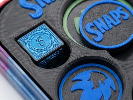 Close-up of a 3D-printed snapboard game tray with blue and black pieces, including a blue numbered token and circular snap pieces.