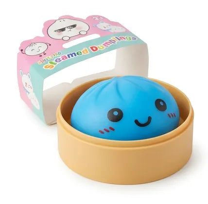 A cute blue steamed dumpling toy with a smiling face, sitting inside a round beige container. The packaging box behind it is pink and light blue with cartoon dumpling illustrations and the text 'EXCLUSIVE Steamed Dumplings'.