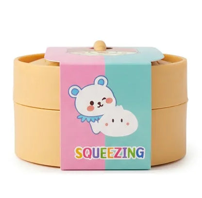 A pastel-colored round container with a lid, wrapped in a pink and light blue paper band featuring a cartoon bear and the word 'SQUEEZING'.